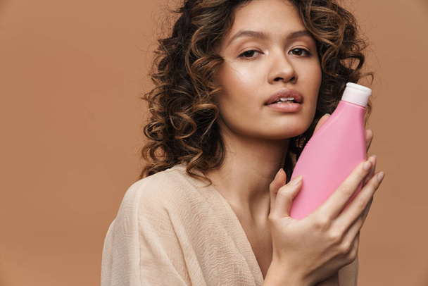 Young hispanic curly woman posing with beauty product isolated over beige background - 写真・画像