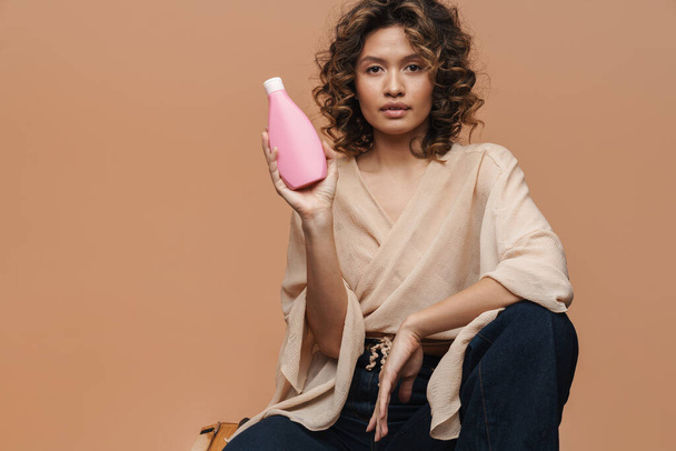 Young curly hispanic woman showing beauty product while sitting isolated over beige background - Φωτογραφία, εικόνα