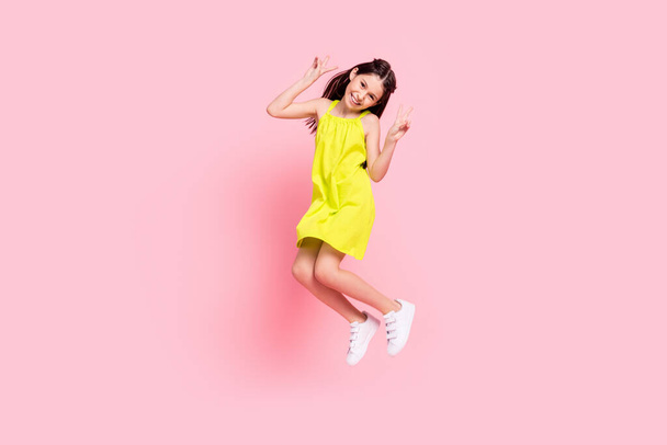Full length photo of pretty funny schoolgirl dressed yellow outfit glasses jumping showing v-sign smiling isolated pink color background - Foto, Imagen