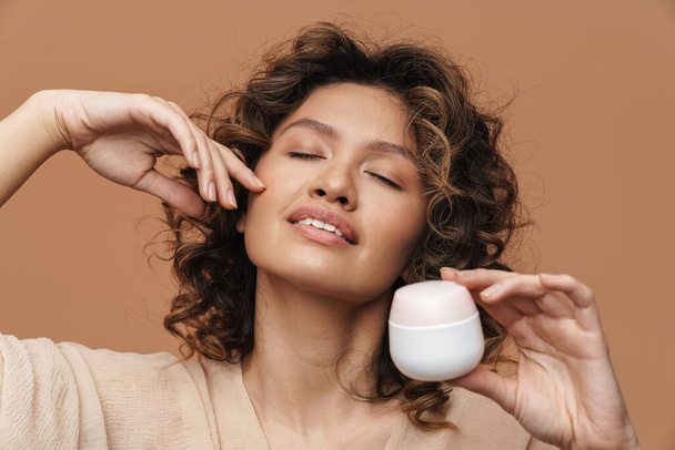 Young curly hispanic woman posing with facial cream isolated over beige background - Φωτογραφία, εικόνα