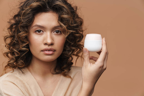 Young curly hispanic woman posing with facial cream isolated over beige background - Foto, Bild