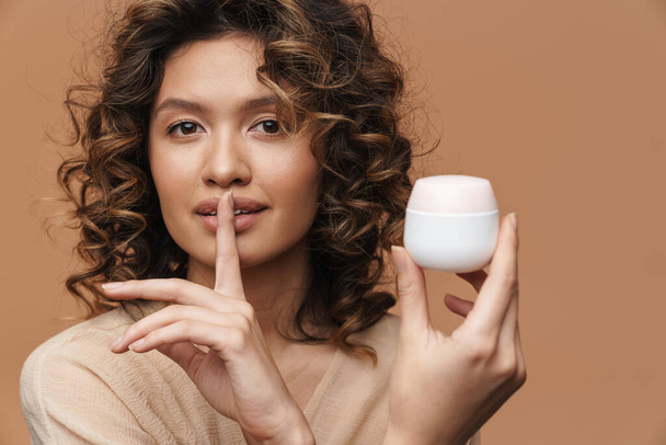 Young woman making silence gesture while showing facial cream isolated over beige background - Foto, afbeelding