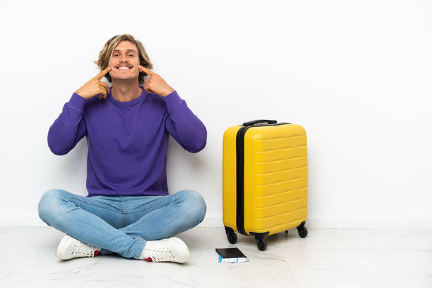 Jeune homme blond avec une valise assise sur le sol souriant avec une expression heureuse et agréable - Photo, image