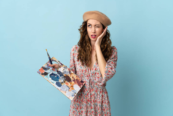 Junges Künstlermädchen hält frustriert eine Palette auf blauem Hintergrund und verdeckt die Ohren - Foto, Bild