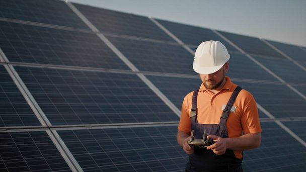 Fotovoltaik güneş enerjisi santralinde kask takan ve insansız hava aracını kontrol eden bir endüstri uzmanı. Güneş paneli kurulumu. Teknoloji ve ekoloji. Yeni teknolojiler - Fotoğraf, Görsel