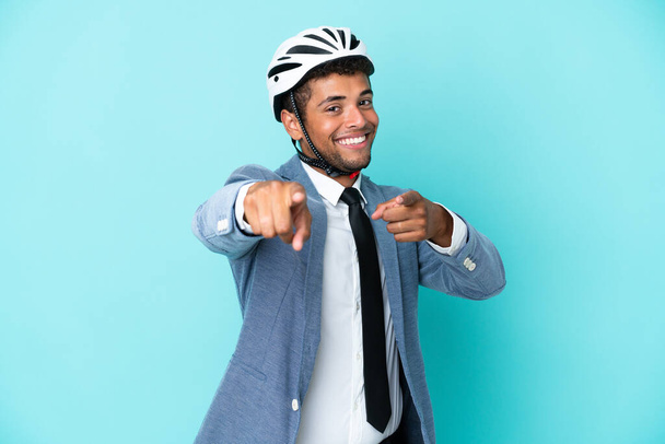 Bisiklet kasklı genç Brezilyalı adam mavi arka planda tek başına sana gülümserken parmak atıyor. - Fotoğraf, Görsel