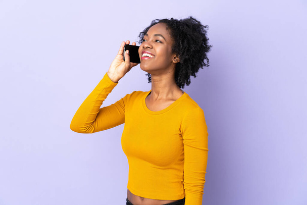 Молодая африканская американка, изолированная на фиолетовом фоне, поддерживает разговор с мобильным телефоном - Фото, изображение