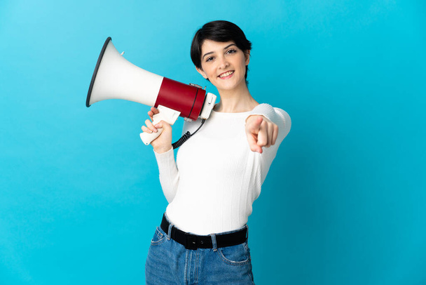 Woman with short hair isolated on blue background holding a megaphone and smiling while pointing to the front - Photo, Image