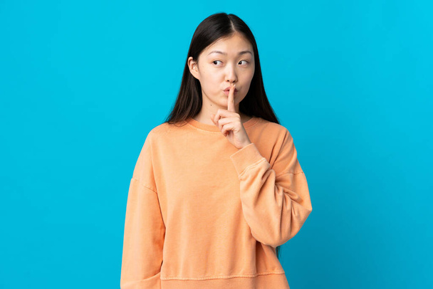 Junges chinesisches Mädchen auf isoliertem blauem Hintergrund, das ein Zeichen der Stille zeigt und den Finger in den Mund legt - Foto, Bild