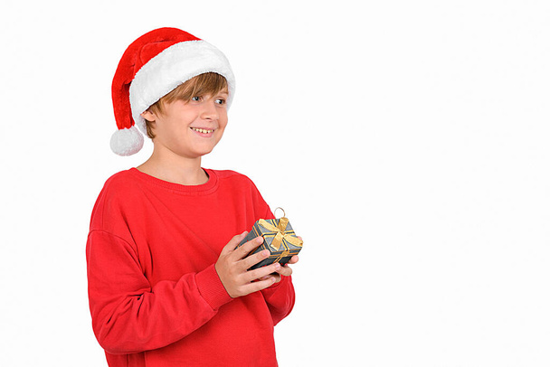Feliz niño rubio sonriente en el sombrero de Santa y jersey rojo, sosteniendo un regalo de Navidad, esperando las vacaciones un nuevo año de ensueño esperando vacaciones de Navidad, esperando regalo y ventas. Feliz año nuevo banner - Foto, Imagen