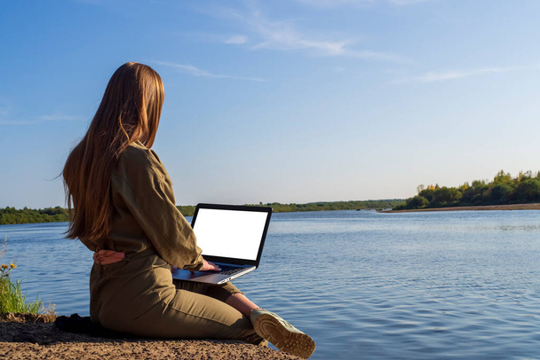 Dziewczyna z laptopem siedzi na brzegu rzeki w parku za miastem. Koncepcja wolnego strzelca, zdalnej pracy lub szkolenia. Niezależna kobieta biznesmen pracuje w przyrodzie podczas podróży. - Zdjęcie, obraz