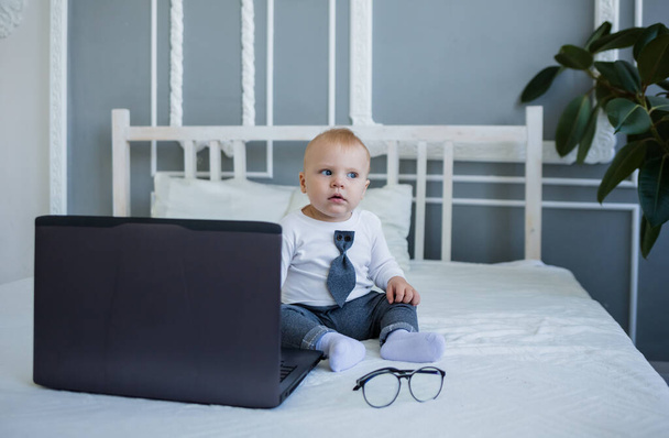 Маленький мальчик в костюме сидит на кровати с ноутбуком - Фото, изображение