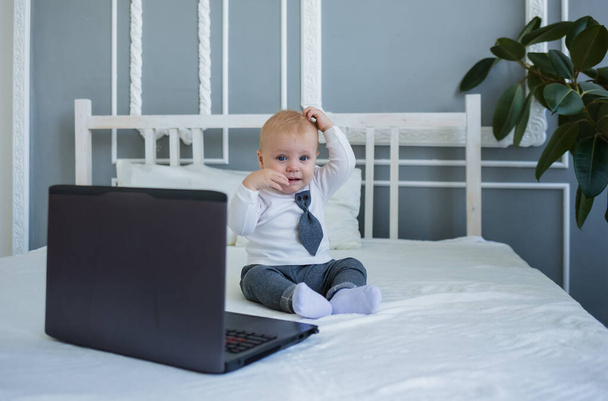Забавный маленький мальчик в костюме сидит на кровати с ноутбуком - Фото, изображение