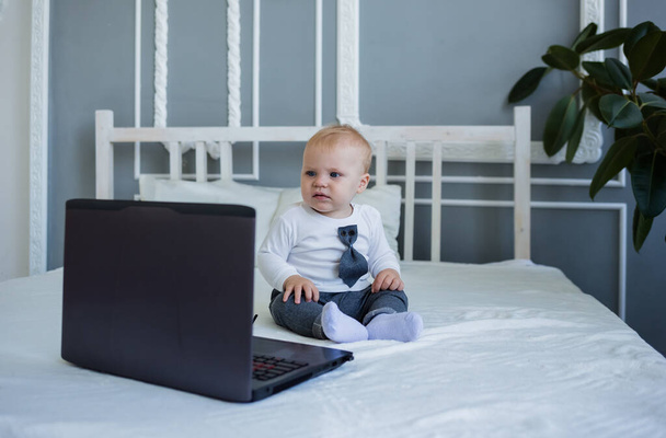 a serious little boy in a suit is sitting on the bed with a laptop - Photo, Image