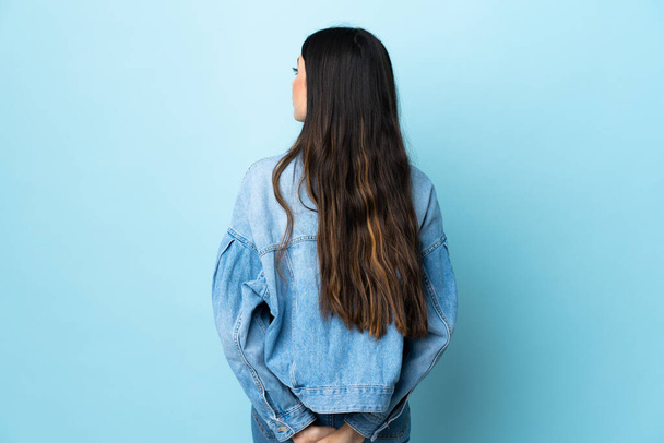 Νεαρή μελαχρινή κοπέλα πάνω από απομονωμένο μπλε φόντο στην πίσω θέση και κοιτάζοντας πίσω - Φωτογραφία, εικόνα
