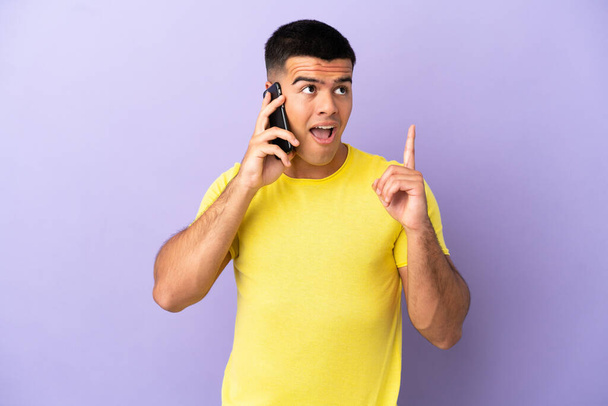 Fiatal jóképű férfi használja a mobiltelefon felett elszigetelt lila háttér gondolkodás egy ötlet rámutatva az ujját fel - Fotó, kép