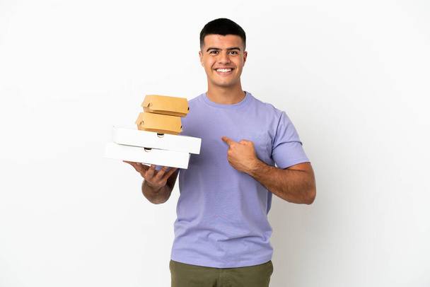 Jeune homme beau tenant des pizzas et des hamburgers sur fond blanc isolé avec expression faciale surprise - Photo, image