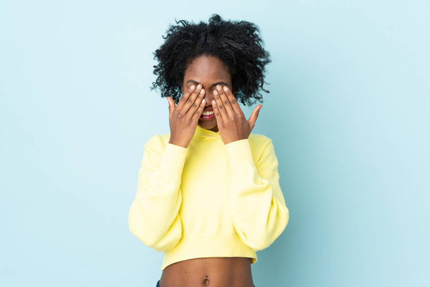 Νεαρή Αφρο-Αμερικανίδα απομονωμένη σε μπλε φόντο που καλύπτει τα μάτια με τα χέρια - Φωτογραφία, εικόνα