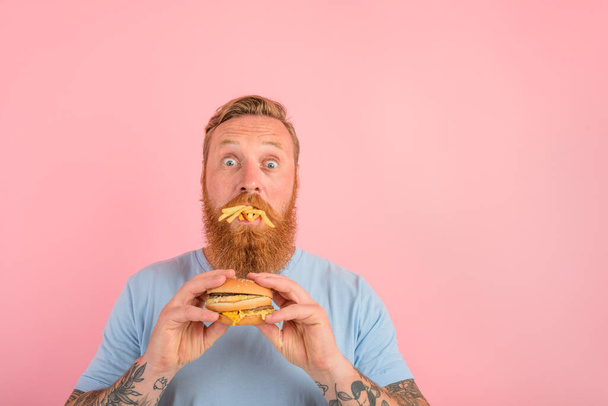 Hämmästynyt mies, jolla on parta ja tatuoinnit, syö voileivän hampurilaisella ja perunoilla. - Valokuva, kuva