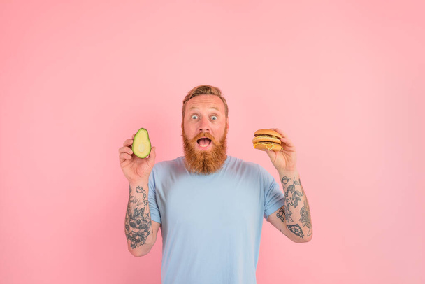 Erstaunter Mann mit Bart und Tätowierungen ist unentschlossen, ob er eine Avocado oder einen Hamburger essen soll - Foto, Bild