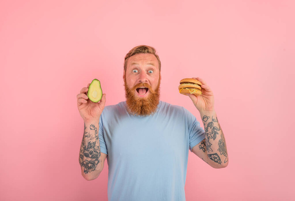 Amazed man with beard and tattoos is undecided if to eat an avocado or an hamburger - Photo, Image