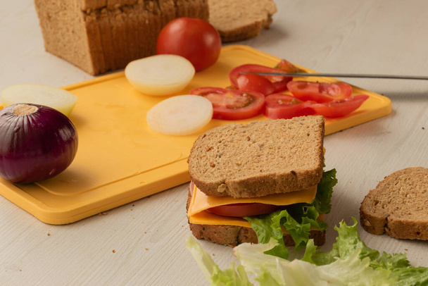 primer plano de delicioso sándwich listo para comer con queso, lechuga, tomate fresco, en una mesa, en el fondo una tabla de cortar con verduras frescas cortadas, aperitivo e ingredientes en el estudio - Foto, imagen