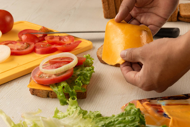 Hände bereiten und legen den Cheddar-Käse eines leckeren Sandwiches mit Salat, frischen Tomaten- und Zwiebelringen auf einen Tisch, im Hintergrund ein Schneidebrett mit frisch geschnittenem Gemüse, Snacks und Zutaten im Studio - Foto, Bild