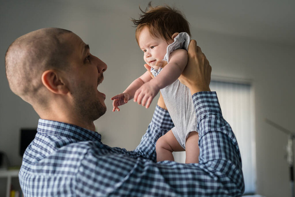 Adult caucasian man father wearing blue shirt holding his small five months old baby girl child while standing by the window at home fatherhood and parenting concept father's day real people side view - Foto, immagini