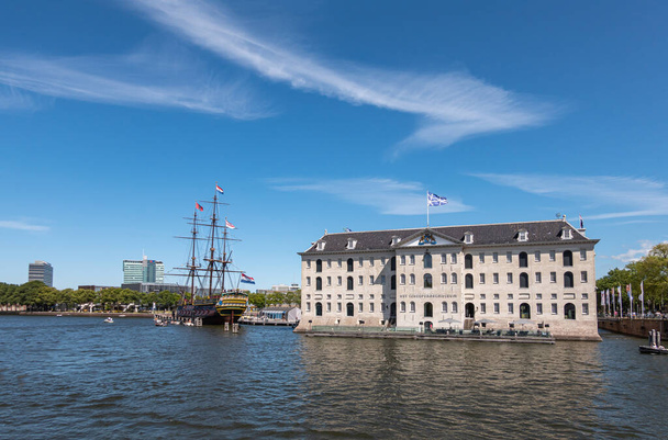 Amsterdam, Paesi Bassi - 15 agosto 2021: Ampio colpo alla pietra beige Edificio del museo marittimo o Scheepvaart e autentico veliero mercantile sotto il cielo blu. - Foto, immagini