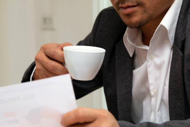 Zblízka mladý asijský podnikatel v černém obleku drží šálek kávy a prodej zpráva papíru myšlení v úřadu - Fotografie, Obrázek
