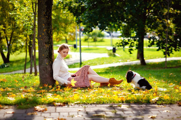 A beautiful blonde woman is playing in the autumn sunny park with her Cavalier King Charles Spaniel dog. A woman and her pet in nature. - Photo, Image