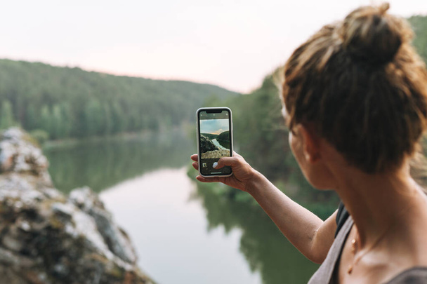携帯電話で山と穏やかな川の美しい景色を撮影バックパックを持つ若いスリムな女性は、地元の旅行 - 写真・画像