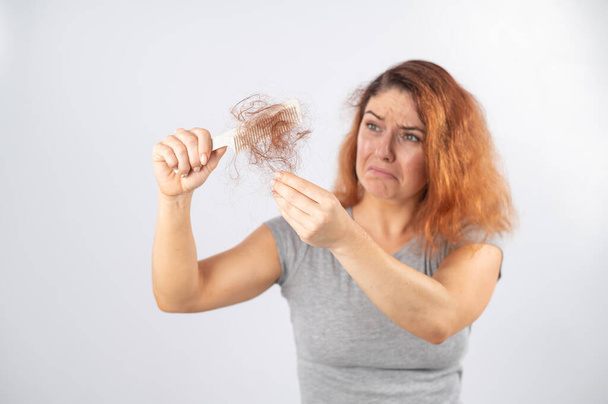 Caucasian woman with a grimace of horror holds a comb with a bun of hair. Hair loss and female alopecia. - Foto, imagen