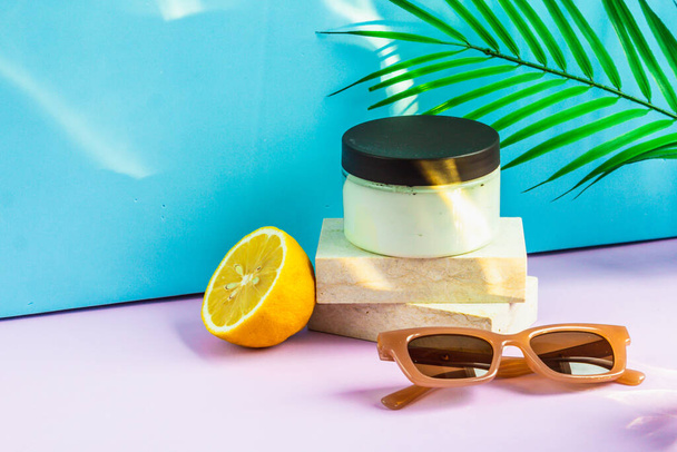 Cosmetics are on the podium on a summer background, in the rays of the sun. a jar of cream is on the podium, half a lemon and sunglasses are lying next to it, and a palm leaf is hanging above it. - Foto, immagini