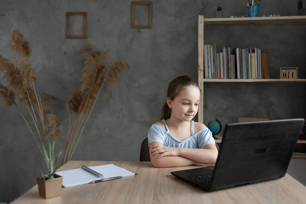 An elementary school student listens to an online lesson via video communication at home at her desk, studying remotely with a laptop or pc. The schoolgirl is in touch with an online language teacher - Photo, Image