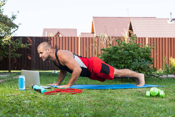 The young man goes in for sports in park. Cheerful sportsman  does push-ups and watching online workout from laptop  in backyard  in summer day - Photo, Image