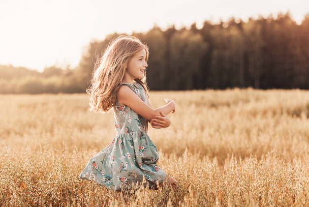 Una niña feliz corre a través de un campo de trigo en el verano en un día soleado. Verano. Vacaciones de verano. Feliz infancia. Emociones positivas y energía - Foto, imagen