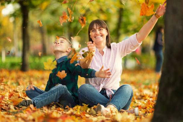 Mom and son are sitting in the park on the ground in bright orange autumn leaves. Happiness, love and good family relationships. Beauty of nature. - Zdjęcie, obraz
