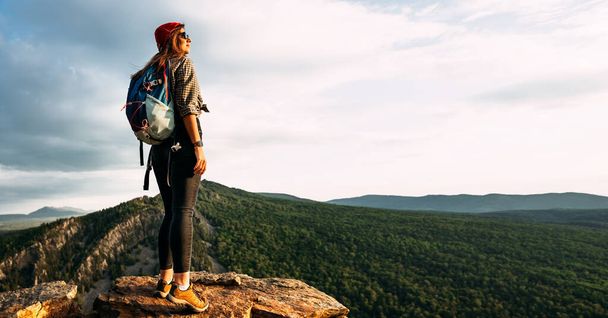 A young female tourist with a backpack admires the sunset from the top of the mountain, panorama. A traveler on the background of mountains. Tourist with a backpack on the background of mountains - Valokuva, kuva
