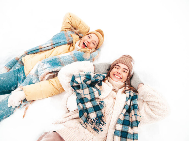Two young beautiful smiling hipster female in trendy warm clothes and scarfs.Carefree women posing in the street in park. Positive pure models lying in snow. Enjoying winter moments. Xmas concept - Zdjęcie, obraz
