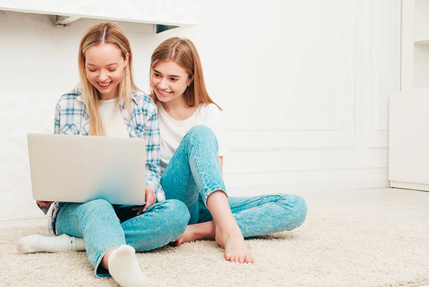 Two young beautiful smiling female in trendy summer clothes. Sexy carefree women posing in light interior in studio. Positive models having fun indoors.Using computer laptop.Looking at notebook screen - Φωτογραφία, εικόνα