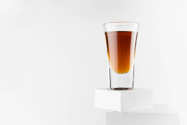 Bright brown brandy in glossy shot glass in sunlight with reflections on abstract white square podiums in soft light space, exquisite minimal style. - Фото, изображение