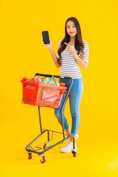 ポートレート美しい若いアジアの女性スーパーマーケットから食料品を買い物し、黄色の隔離された背景にスマートフォンでカート - 写真・画像