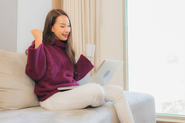 肖像美しい若いアジアの女性はリビングルームのインテリアでソファの上にコンピュータのラップトップを使用します - 写真・画像
