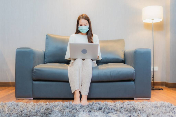 Retrato hermosa joven mujer asiática usar máscara y el ordenador portátil en el sofá en el interior de la sala de estar - Foto, Imagen