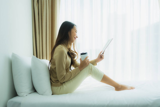 Joven mujer asiática con taza de café y leer libro en la cama en el interior del dormitorio - Foto, imagen