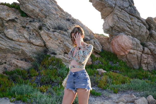 Outdoor portrait of young fashionable woman in crop top and denim shorts with rock formation in background - Photo, Image