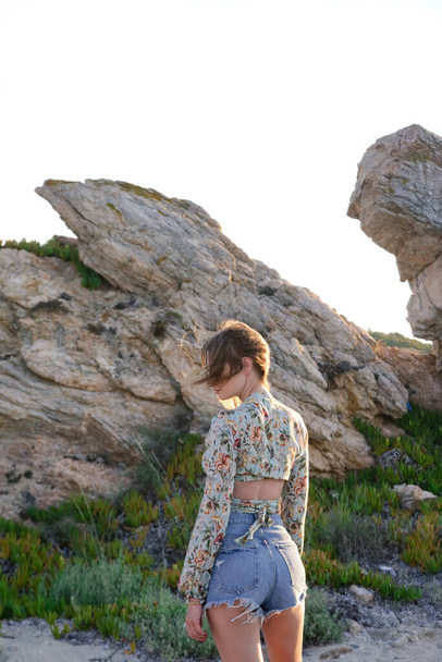 Outdoor-Porträt einer jungen modischen Frau in bauchfreiem Top und Jeans-Shorts mit Felsformation im Hintergrund - Foto, Bild