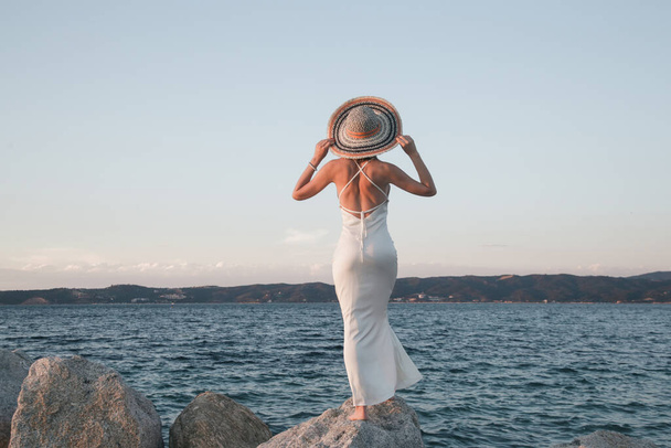 Young woman in long white dress and straw hat on the beach. Stylish summer vacation fashion. - Photo, Image