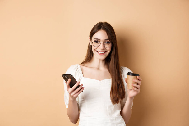 Щаслива біла жінка в окулярах, що стоїть з чашкою кави і смартфона, стоїть на бежевому фоні
 - Фото, зображення